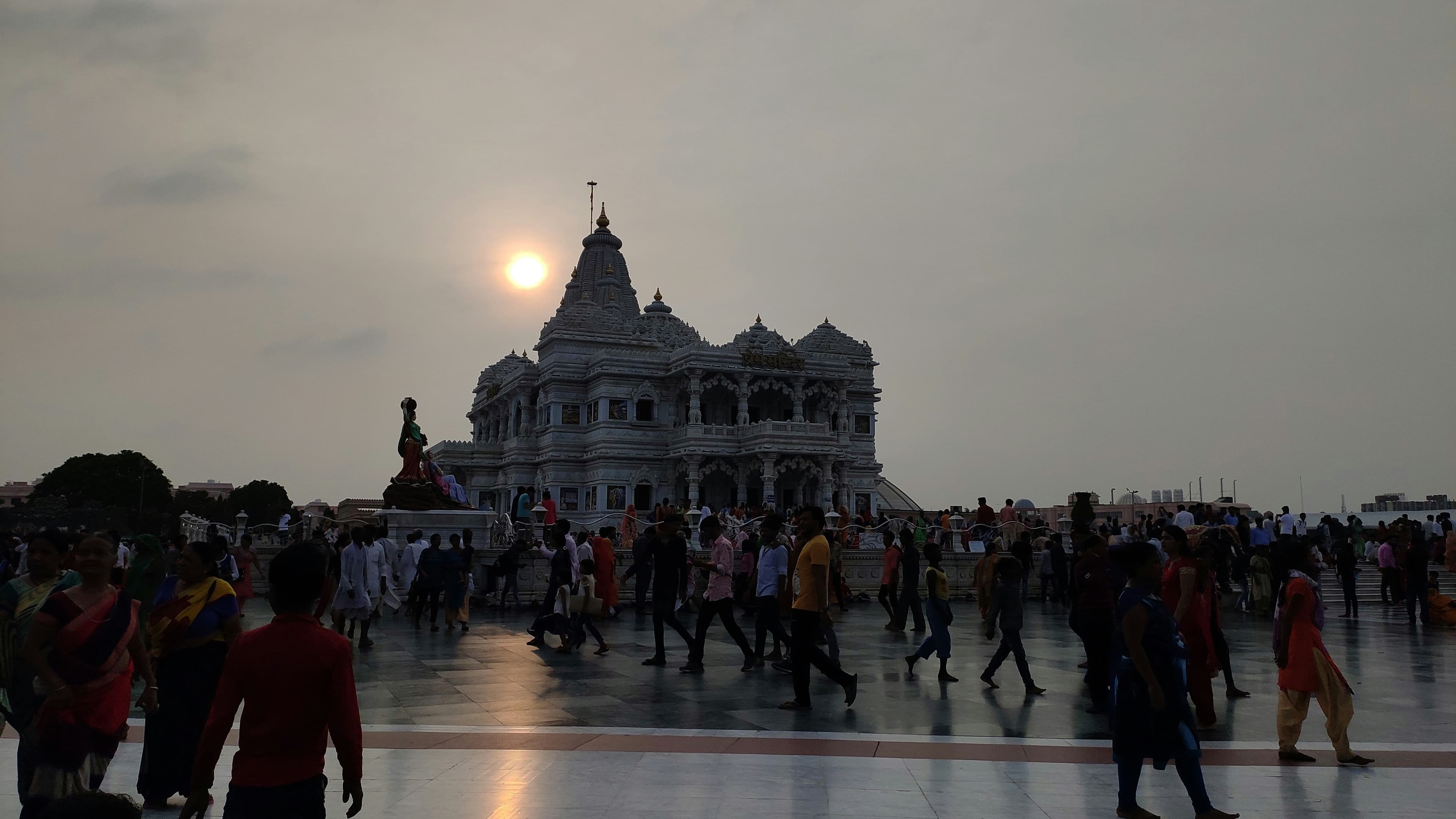 Crowds gather in a reflective courtyard before an ornate white temple at sunset.