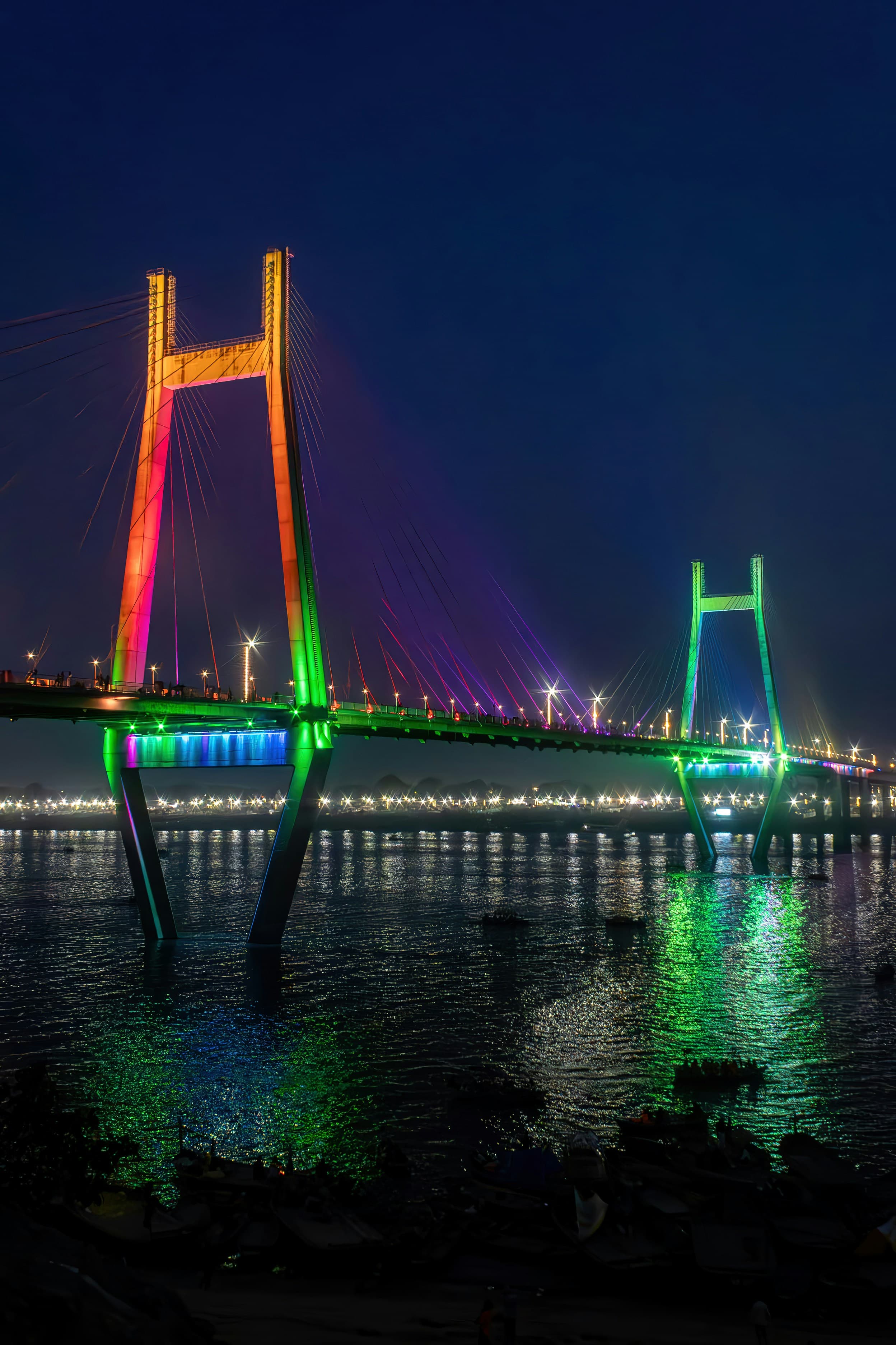 Cable-stayed bridge illuminated with vibrant rainbow colors at night, reflecting on the dark water.